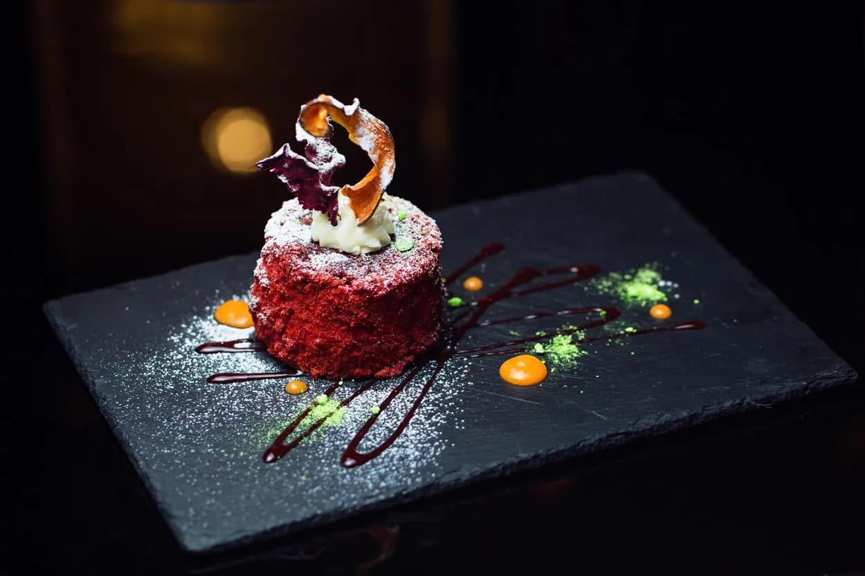 Red velvet cake with cream topping, powdered sugar, and sauces on a dark slate platter