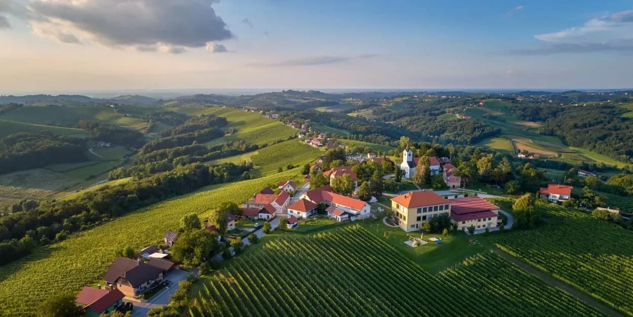 Village nestled among rolling green hills and vineyards under a partly cloudy sky, likely Slovenia.