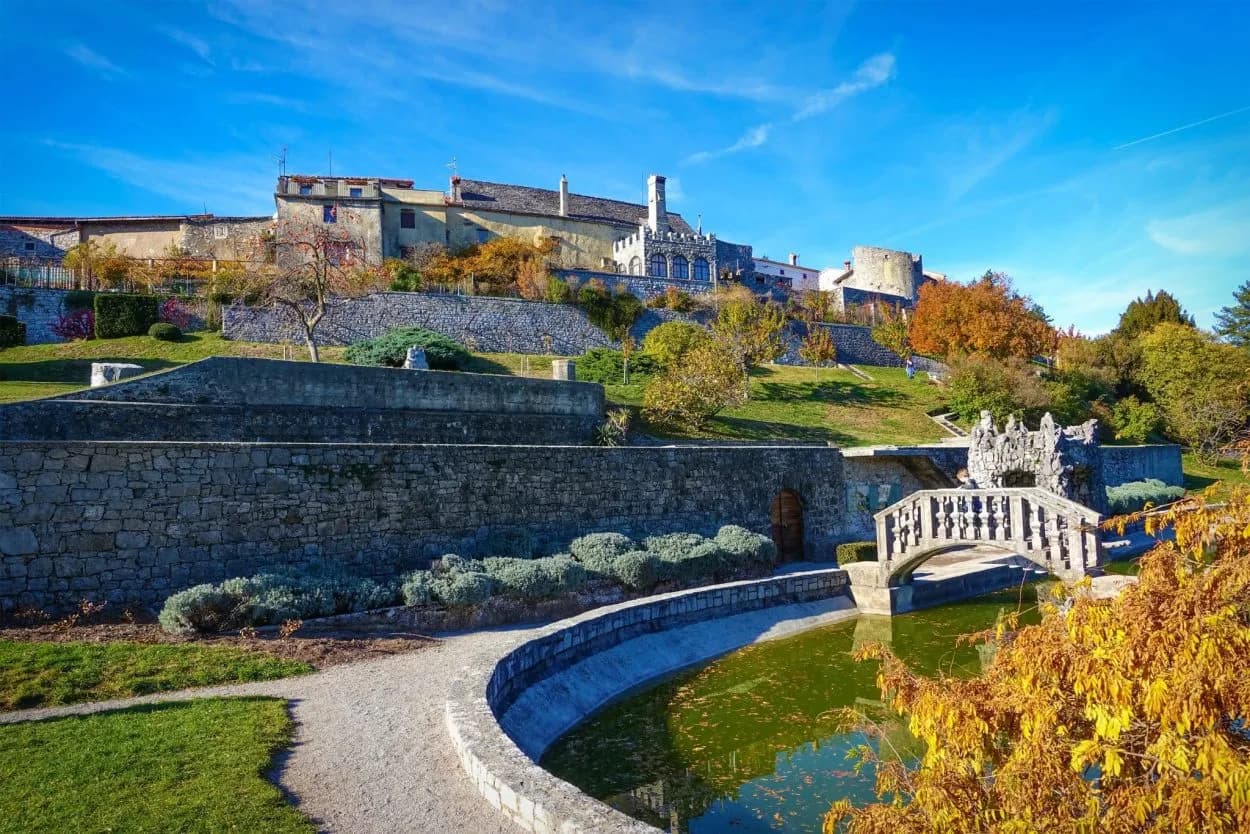 Stone retaining walls and ornate bridge over dark water below hilltop historic buildings in autumn.