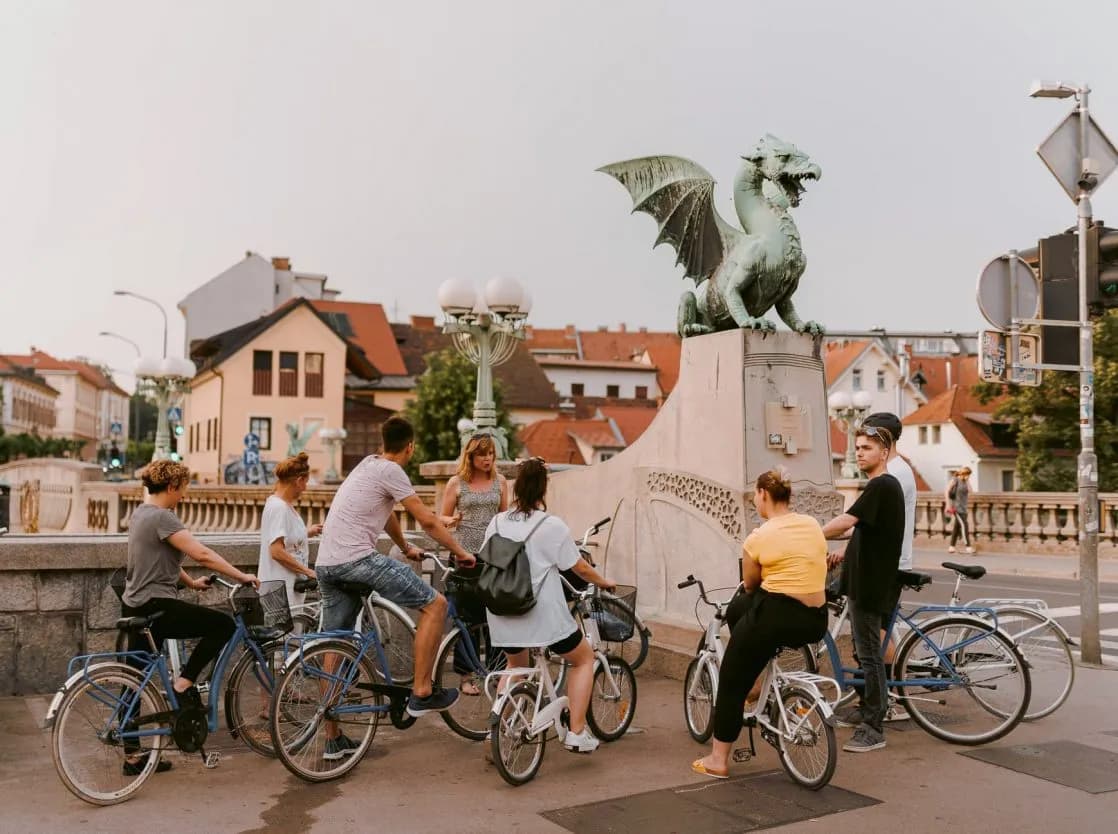 Cyclists gathered near the Dragon Bridge statue in Ljubljana with city buildings in the background.