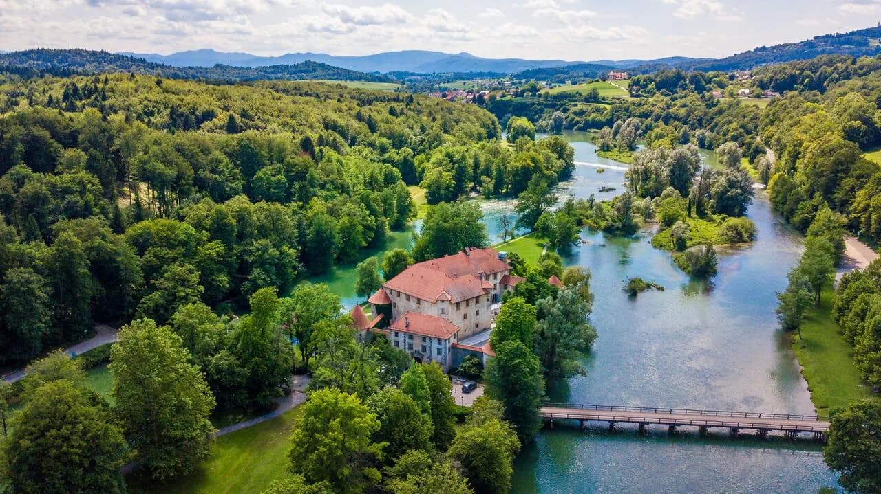 Aerial view of Hotel Grad Otočec with red roof surrounded by dense green forest and a river.