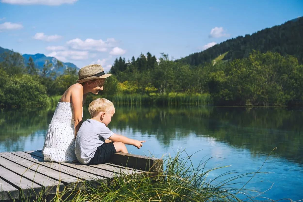 Woman and child on wooden dock pointing at calm alpine lake with forested mountains.