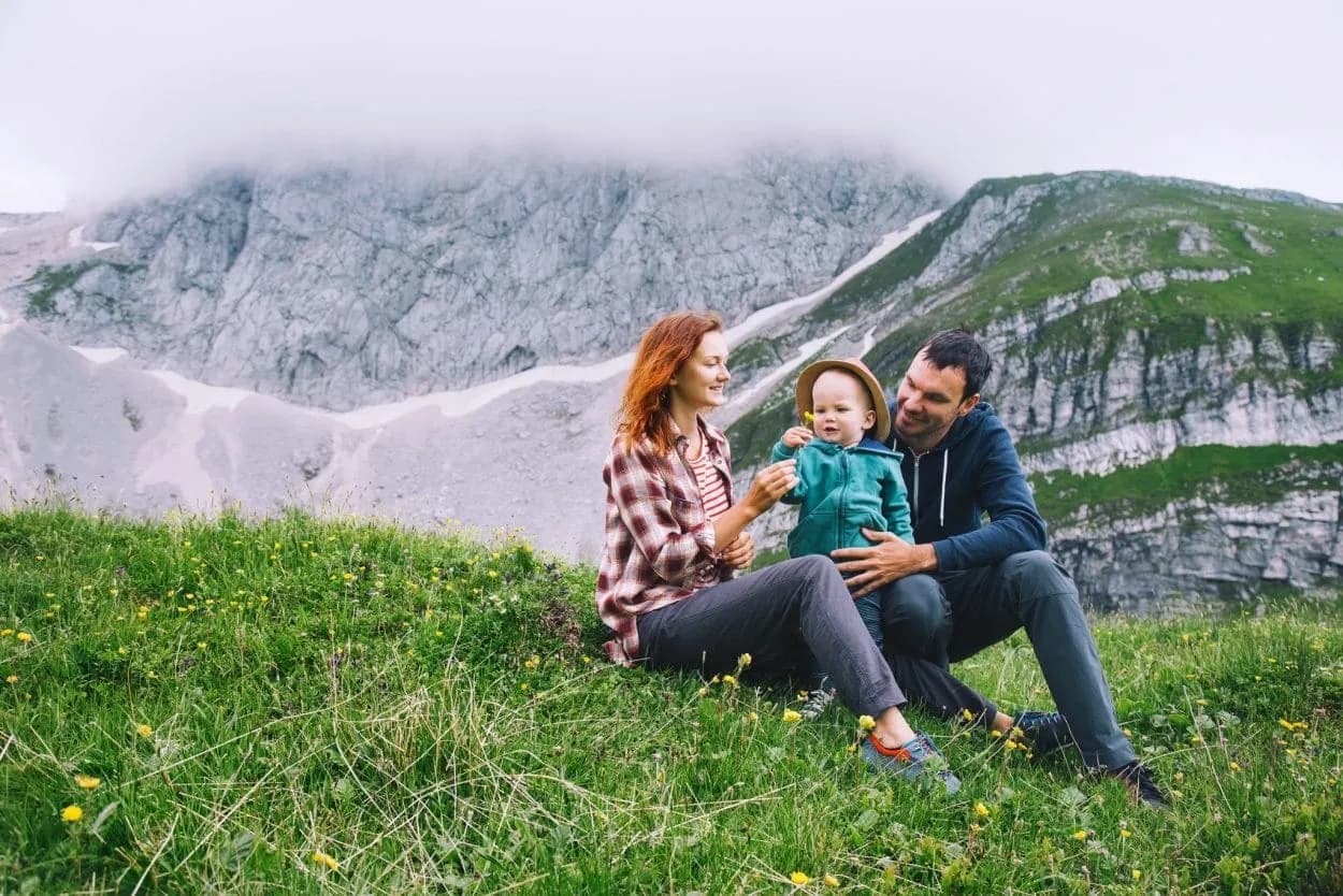 Family relaxing on grassy slope with wildflowers below misty, rocky alpine mountains.