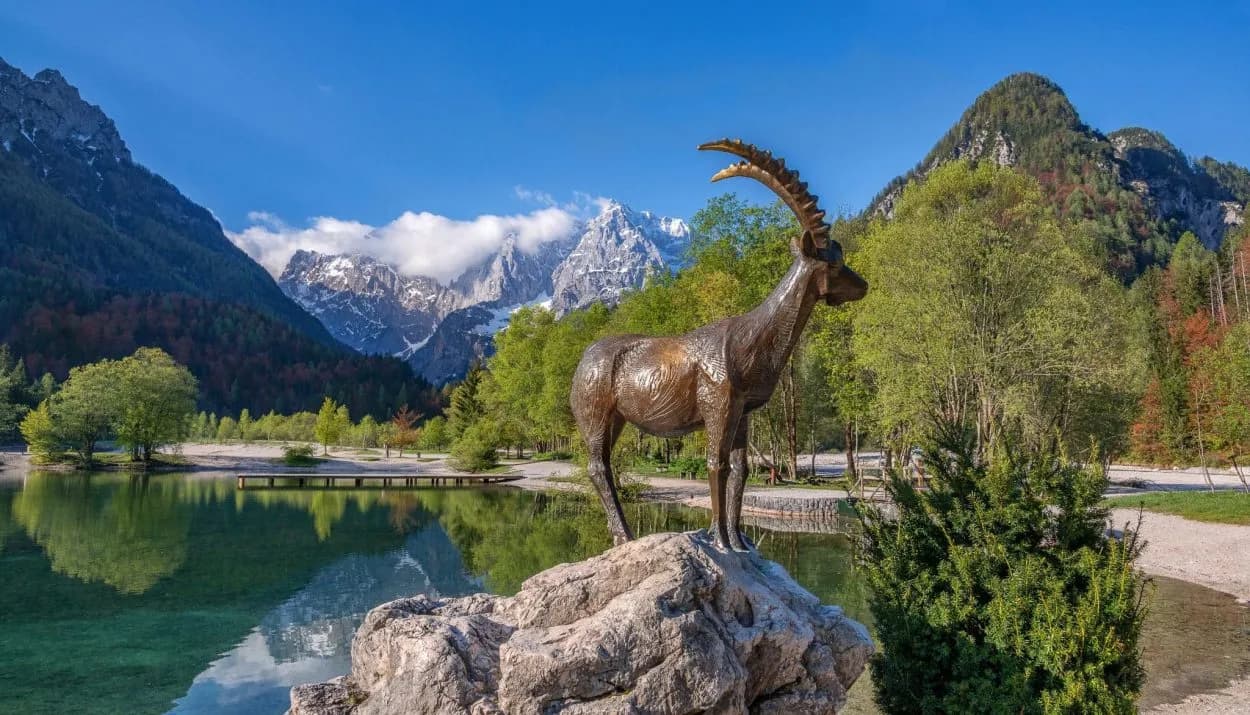 Ibex statue by alpine lake with green trees and snow-capped mountains in Kranjska Gora.