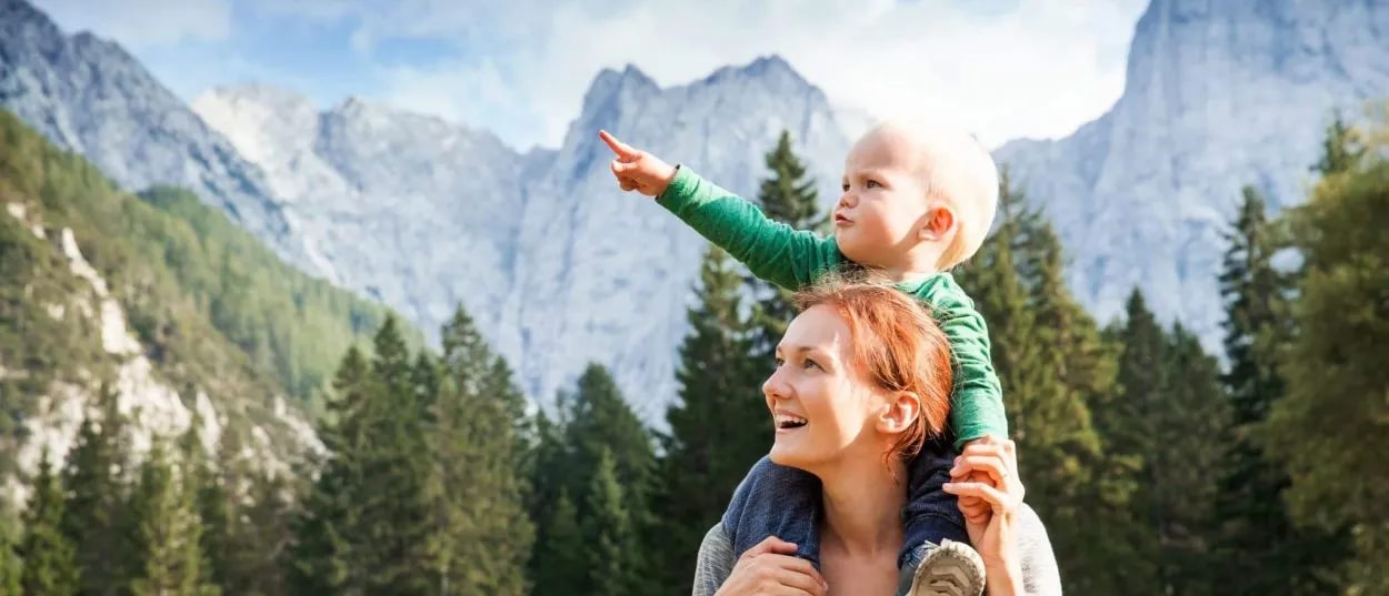 Mother carrying child pointing while hiking near rocky mountains and pine forest in Kranjska Gora.