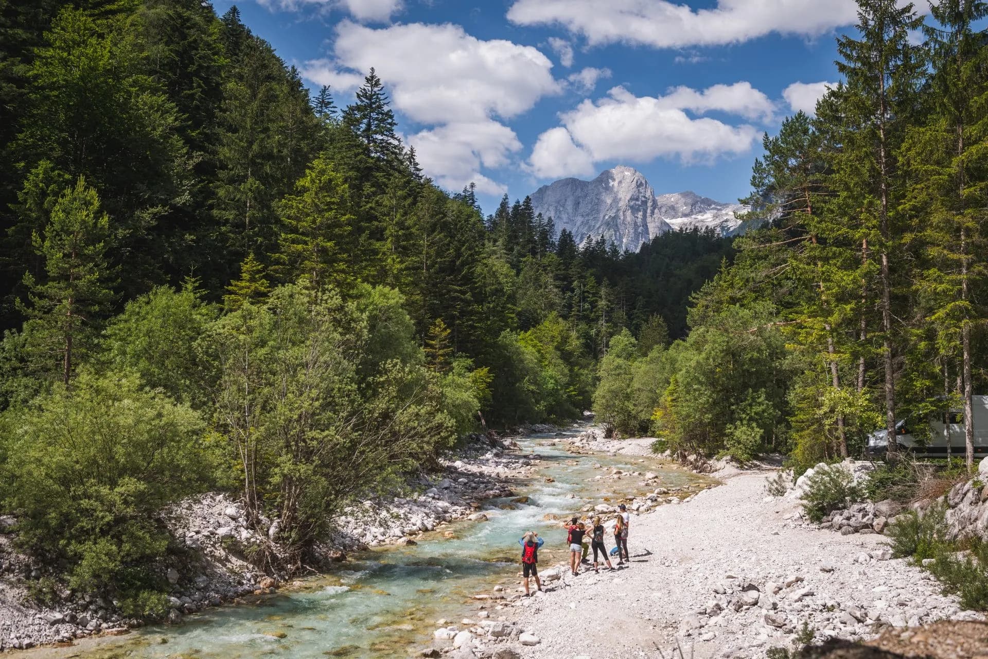 Hikers by clear river in Triglavska Bistrica, Vrata Valley, with forested slopes and rocky mountain peak.