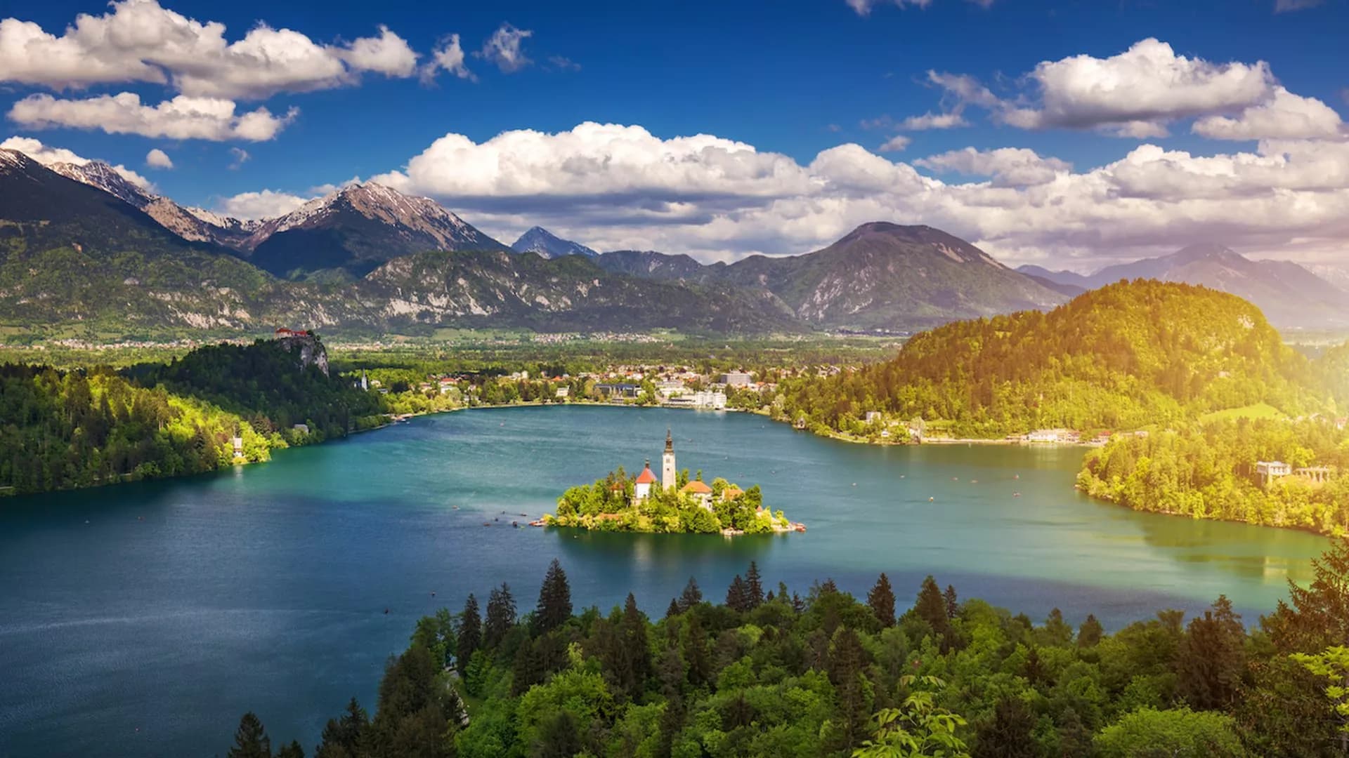 Panoramic view of Lake Bled island church surrounded by mountains and blue sky with clouds.