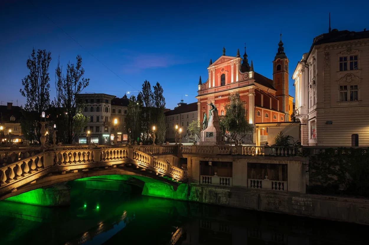 Triple Bridge and pink Franciscan Church illuminated at night in Ljubljana Preseren Square.