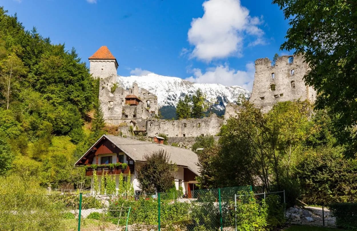 Ruins of Begunje Castle above a house with snow-capped mountains in the background.