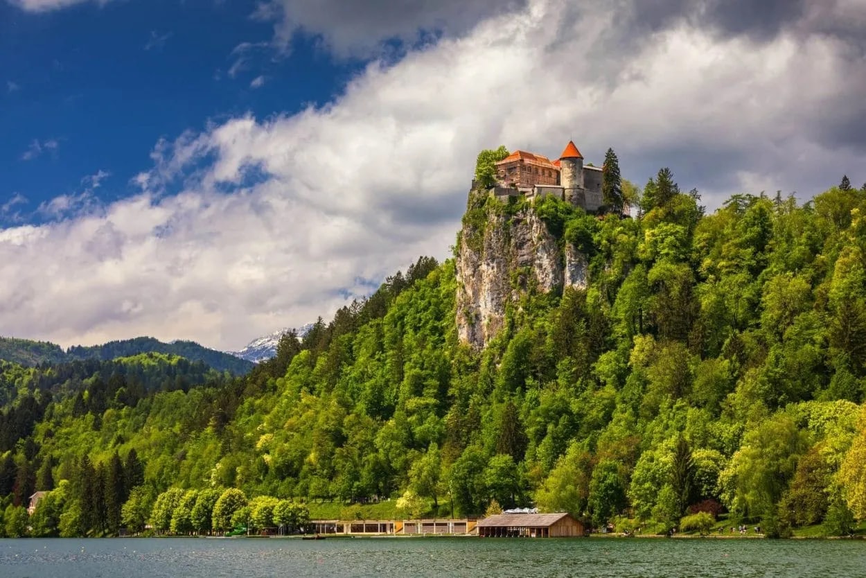 Castle on Bled cliff above lake surrounded by lush green forest under cloudy sky