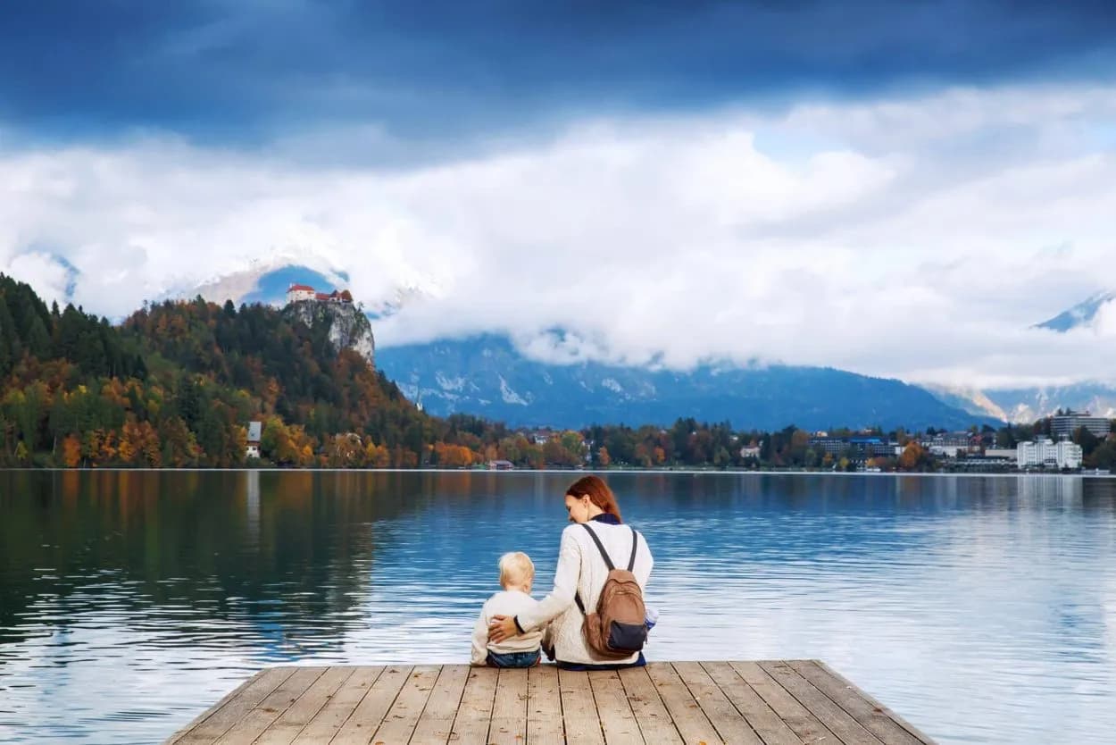 Woman and child sitting on wooden dock overlooking Lake Bled with castle on cliff and mountains