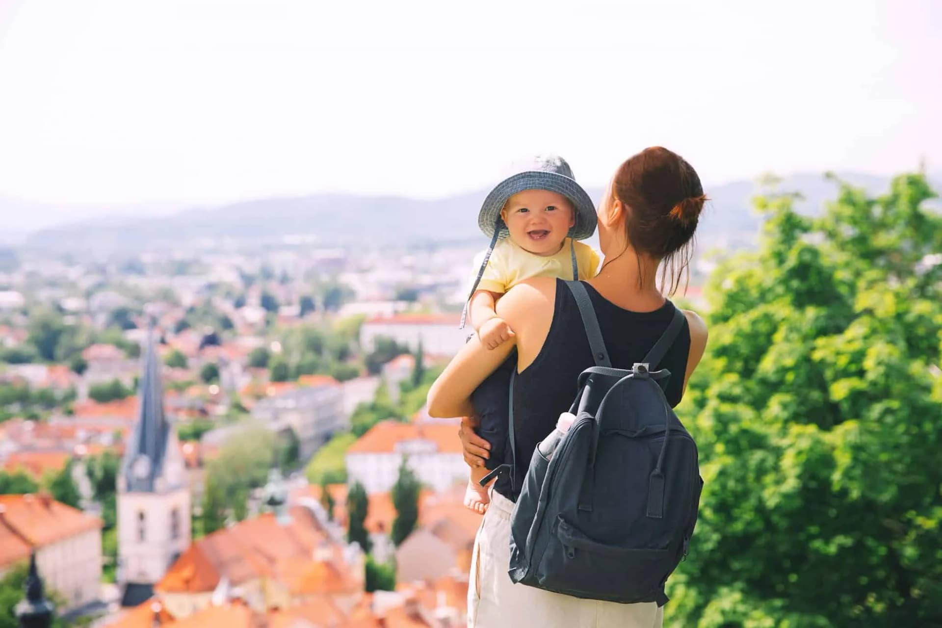Woman holding smiling baby with backpack overlooking Ljubljana's cityscape and church spire.