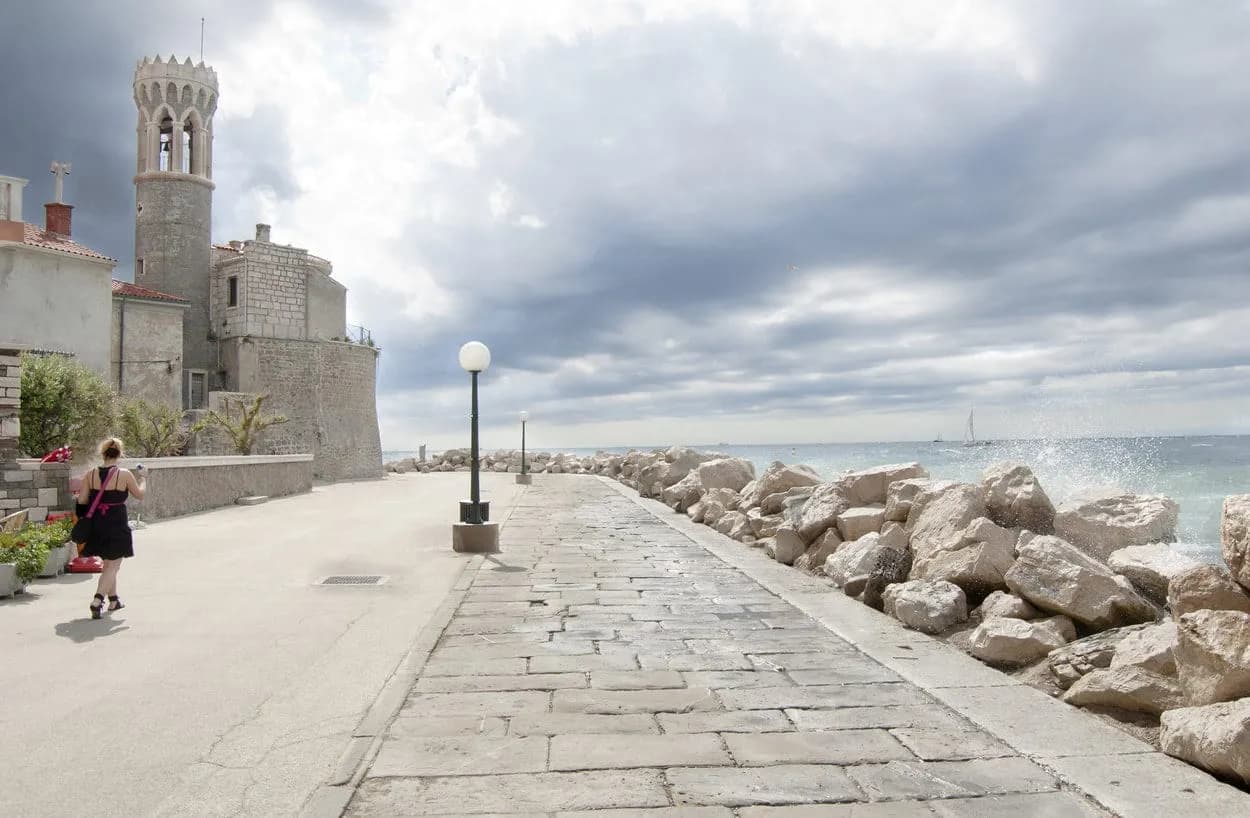 Woman walking by stone seawall with waves crashing, Piran bell tower in background.
