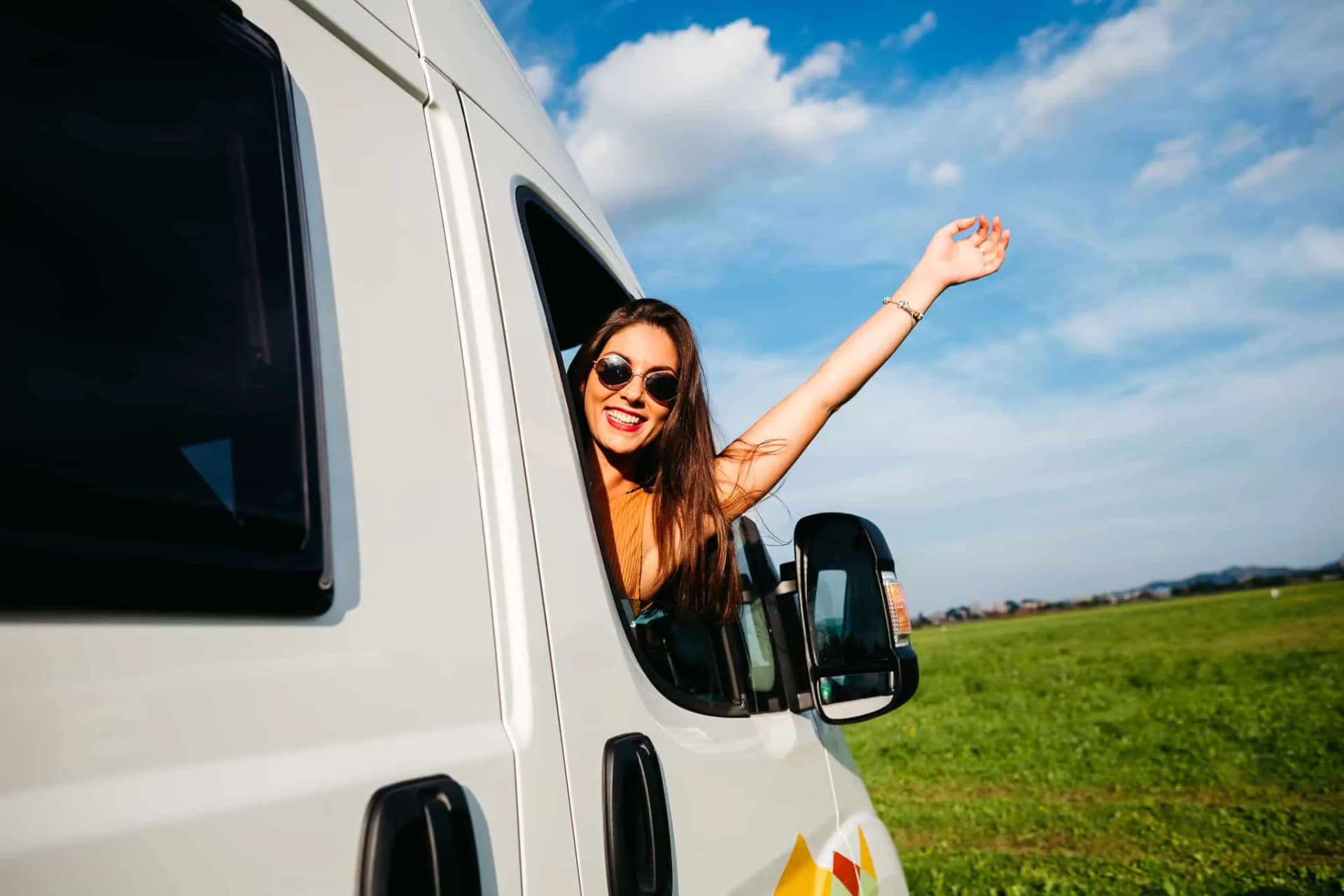Woman leaning out of campervan window with arm raised over grassy field and blue sky
