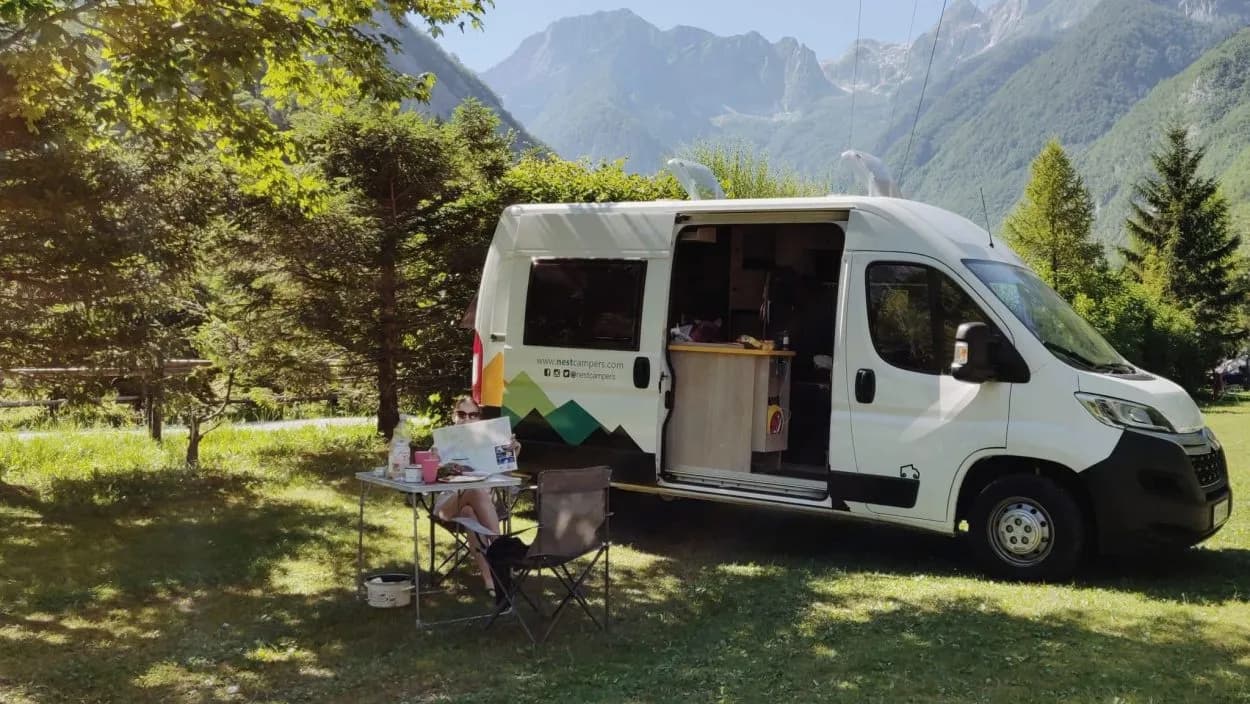 Camper van parked in grassy area with person sitting outside near tall mountains.