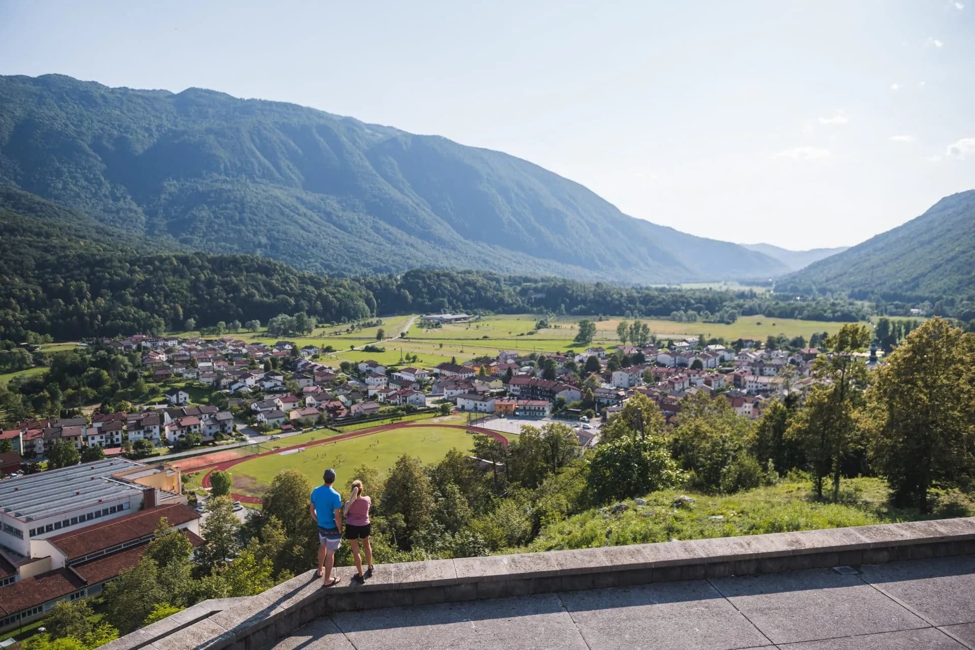 Couple viewing Kobarid town and valley surrounded by steep, forested mountains.