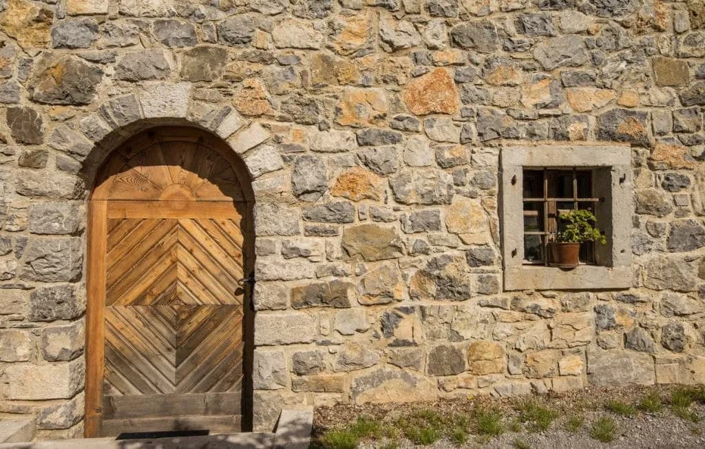 Stone house wall with arched wooden door and small window featuring a potted plant
