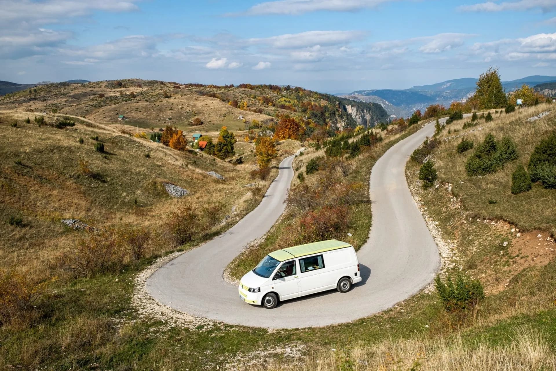 White camper van driving on winding mountain road with dry grass and autumn trees.
