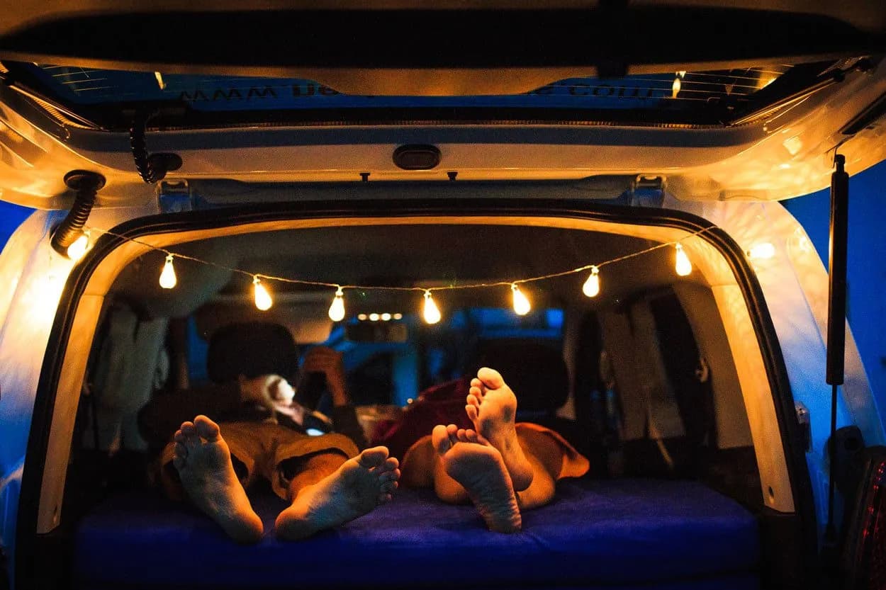 Feet resting in the back of a campervan at night, illuminated by string lights.