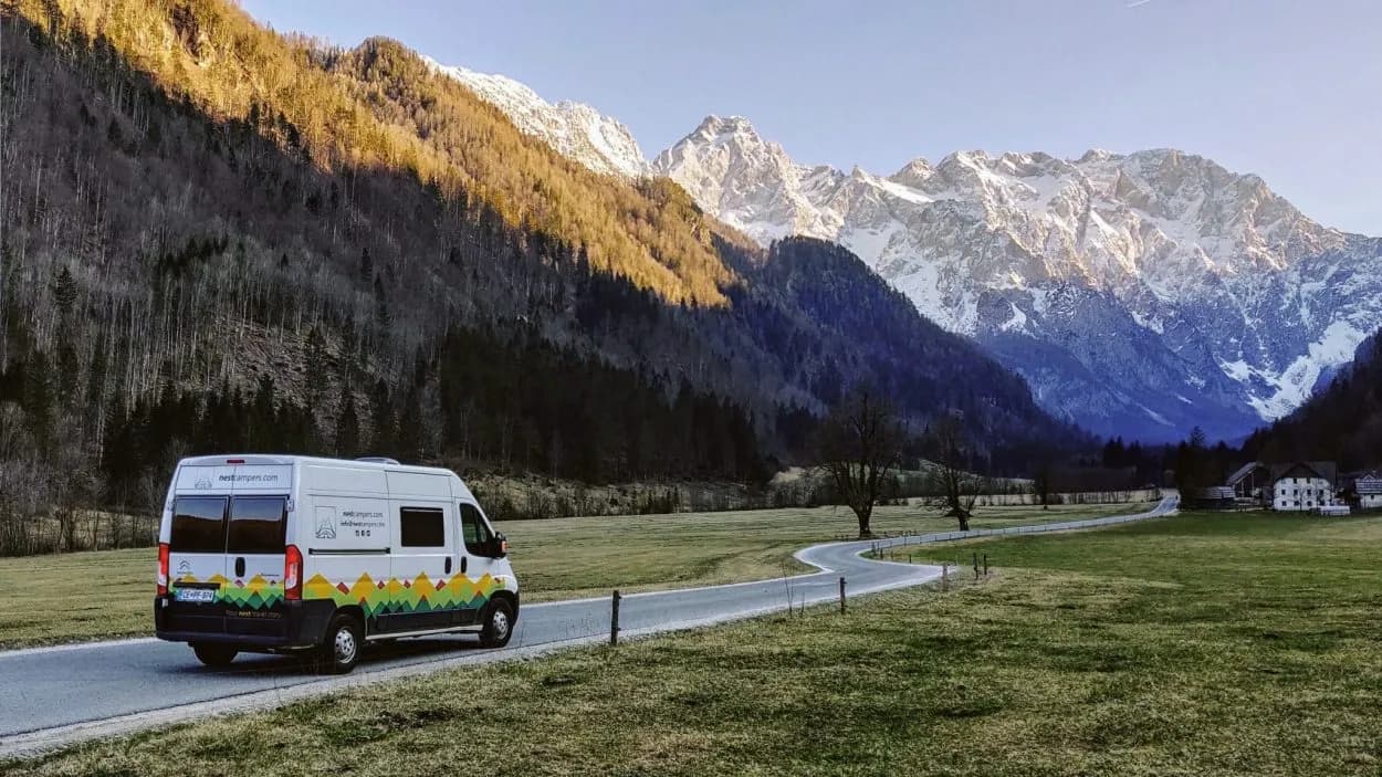 Camper van driving on road through green valley toward snow-capped mountains