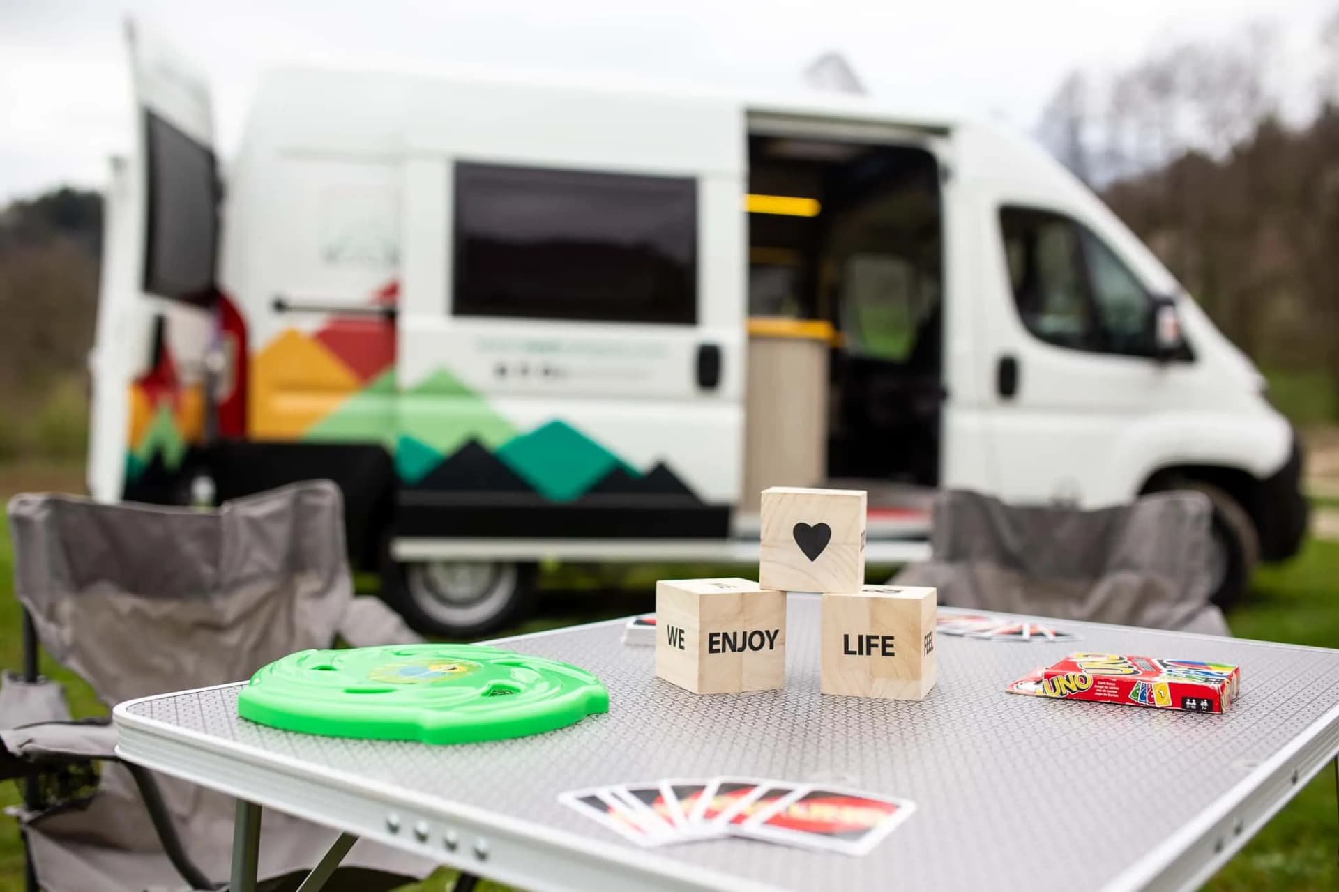 Camping setup with folding table, UNO cards, and wooden blocks outside a camper van.