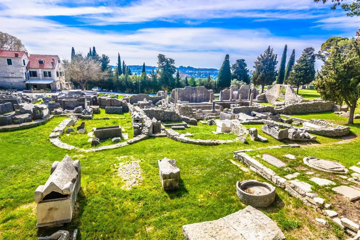Ancient Roman ruins with stone sarcophagi on green grass under a blue sky above Split.