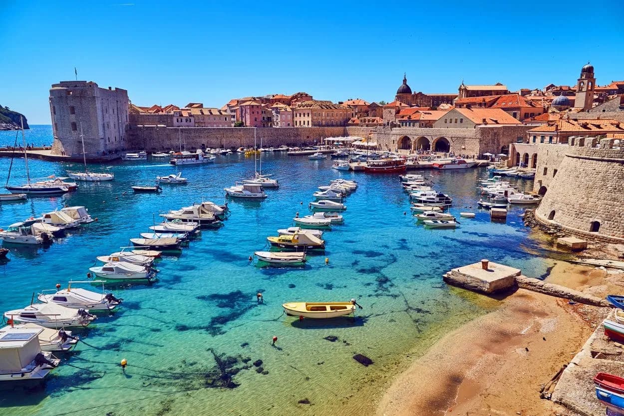 Boats moored in clear turquoise water of Dubrovnik harbor beside historic stone walls.