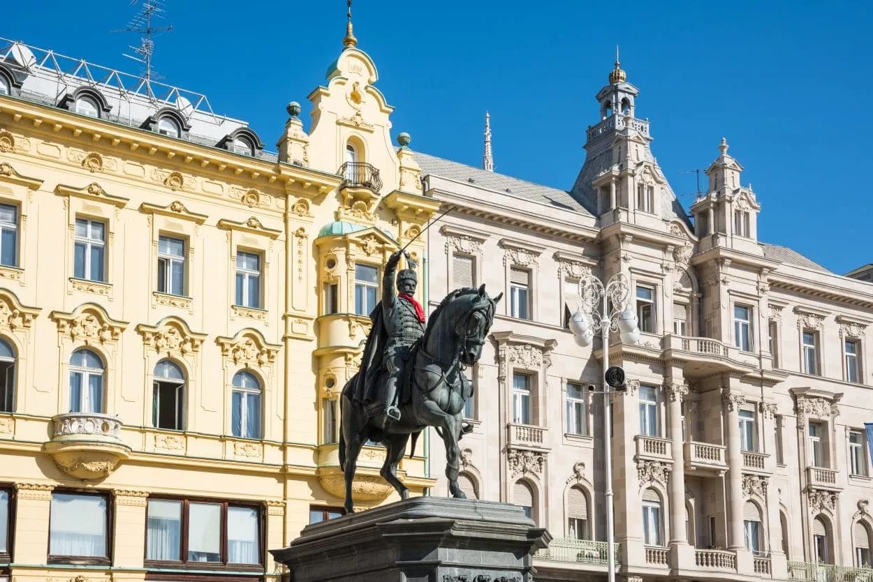 Equestrian statue in Zagreb main square before ornate yellow and white historic buildings under blue sky.