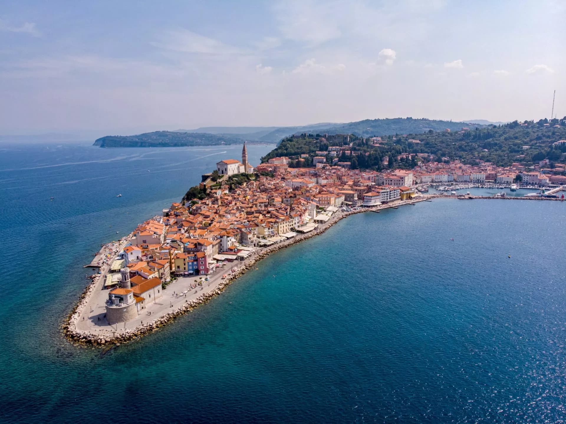 Drone view of Piran, Slovenia coastal town with terracotta roofs and clear blue sea.