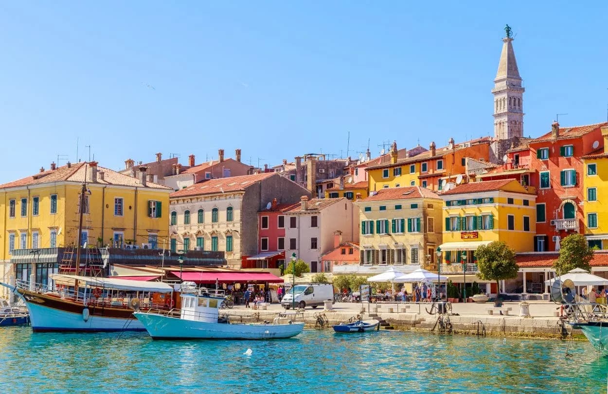 Boats docked in harbor before colorful buildings with a tall bell tower in Rovinj Riviera.