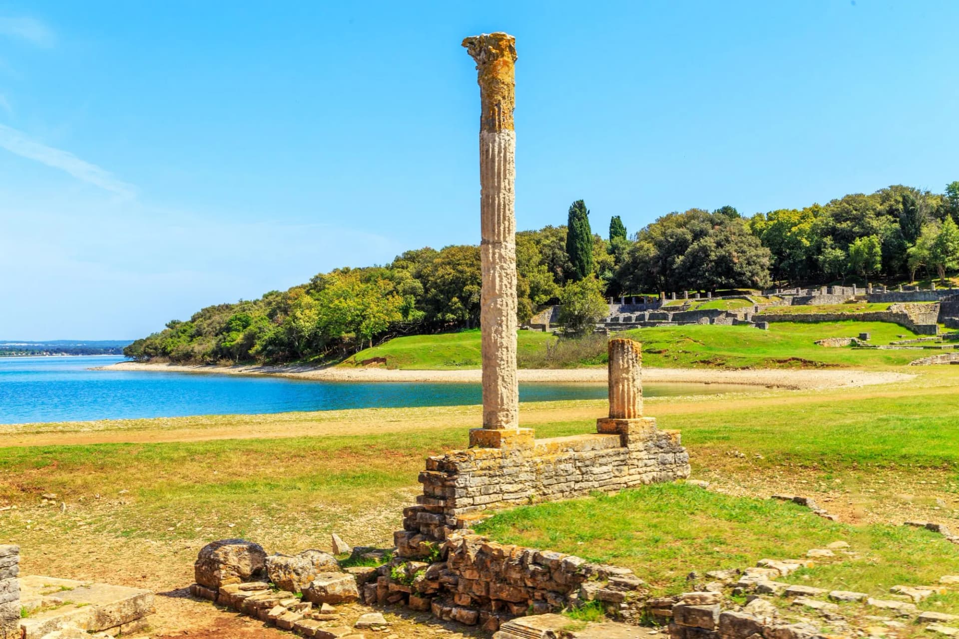 Roman ruins with a tall column beside blue water and green forested coastline in Brijuni National Park.