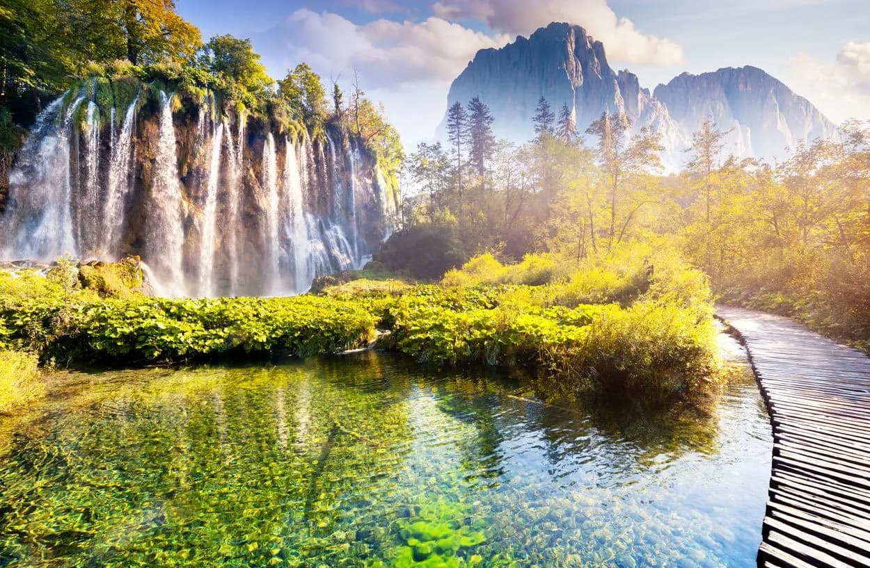 Plitvice Lakes waterfalls, clear green water, wooden boardwalk, and mountain backdrop.