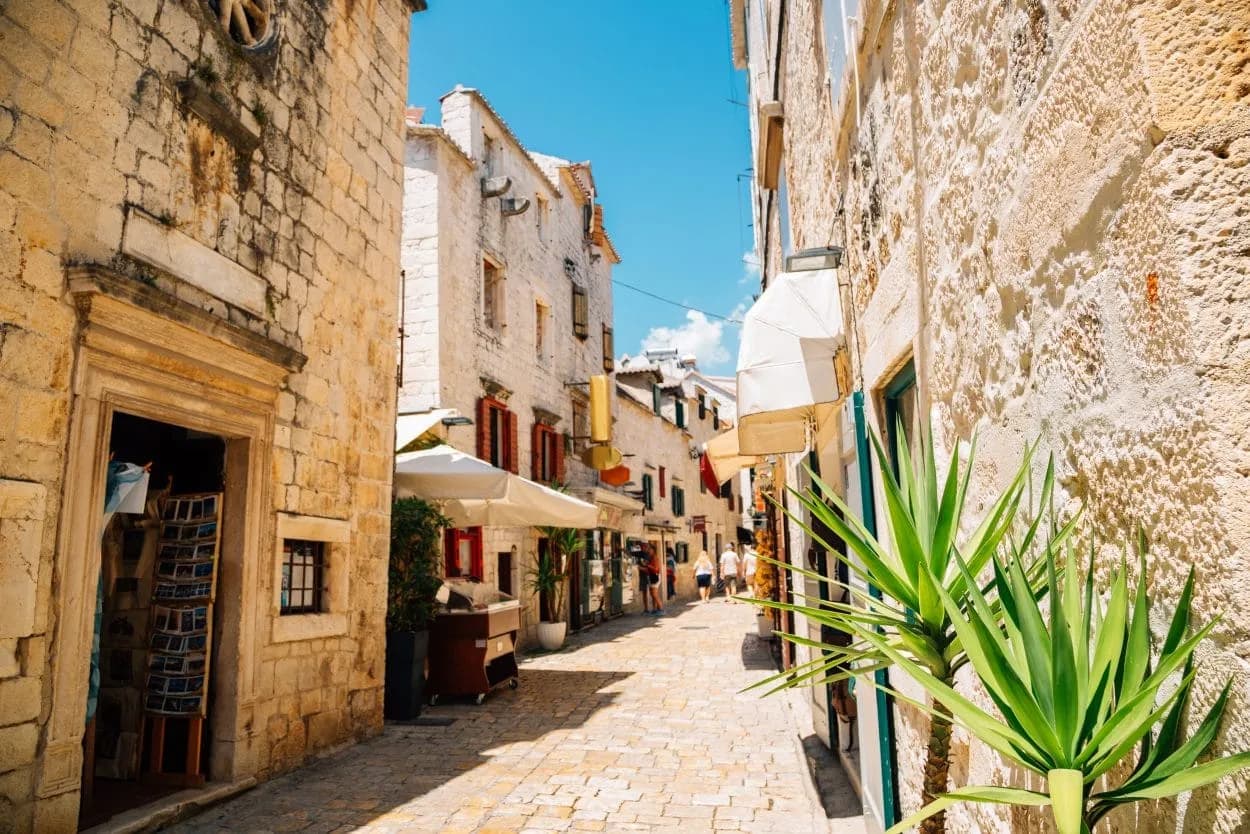 Narrow cobblestone street in Trogir with stone buildings and bright green plants under a blue sky.
