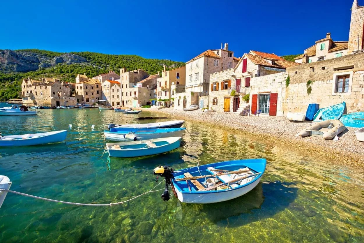 Small blue boats float in clear water near a stone village on Vis island under a bright blue sky.