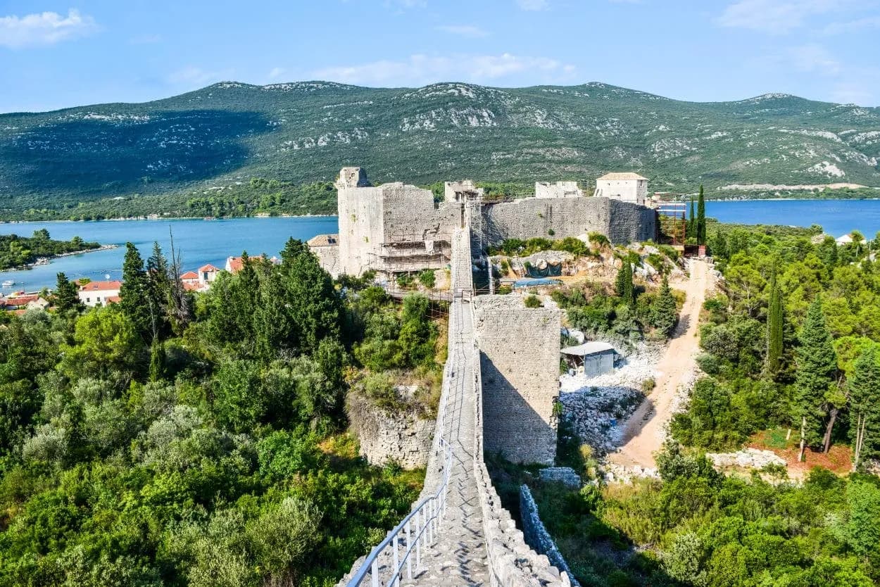 Stone walls of Ston fortress with bridge, lush greenery, and blue water under mountains.