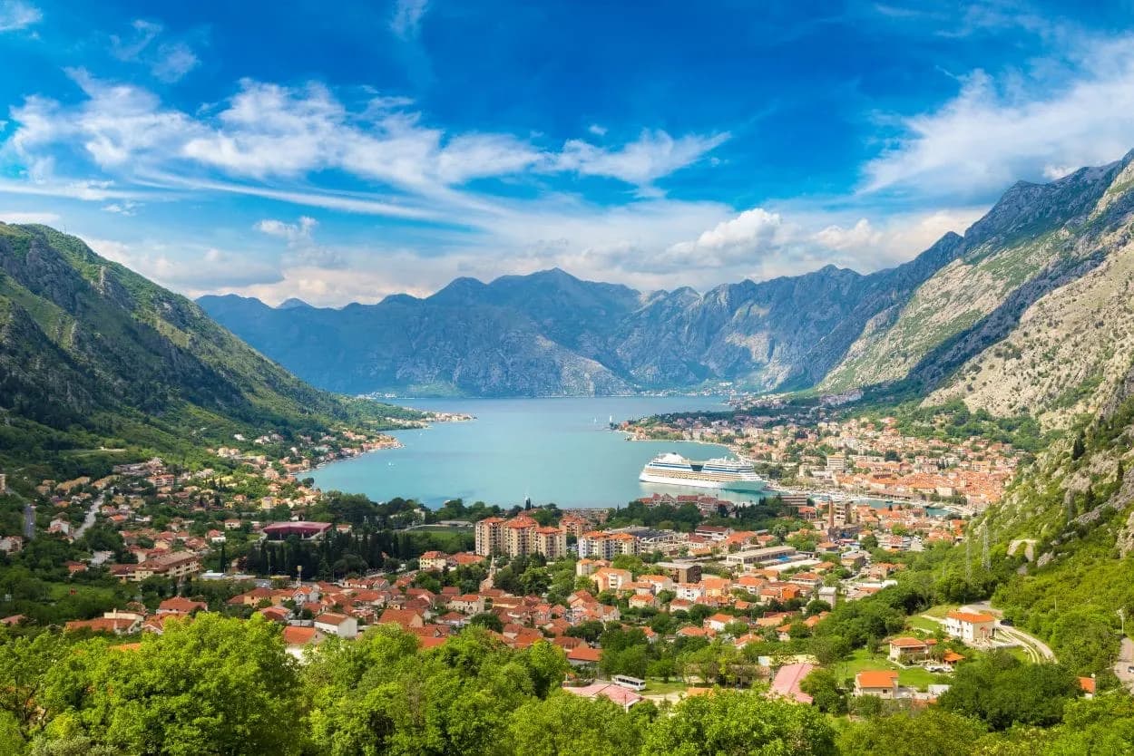 Bay of Kotor panorama with cruise ship, town, and steep green mountains under blue sky.