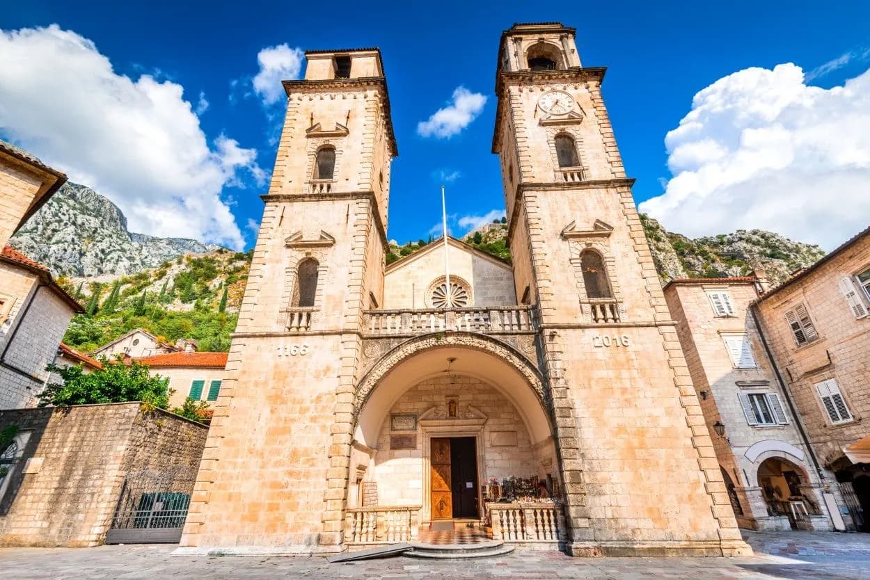 St Tryphon Cathedral with twin bell towers in Kotor, Montenegro, against mountains and blue sky.