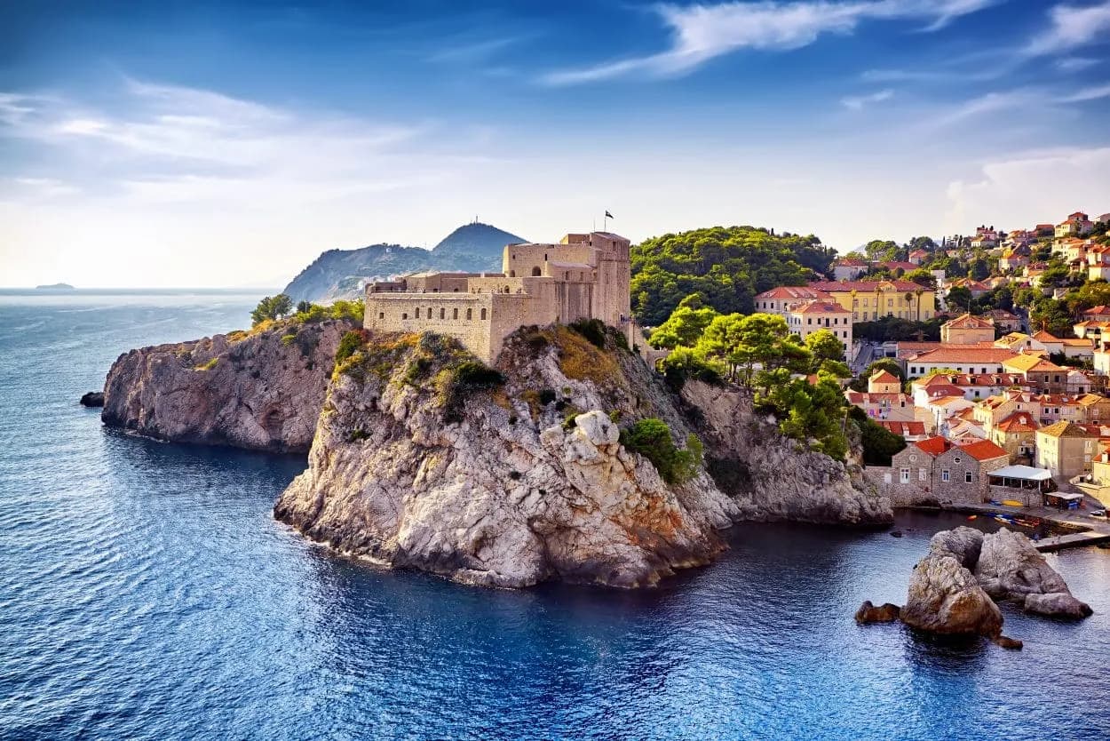Stone fortress on cliff overlooking deep blue sea next to Dubrovnik city with red roofs.