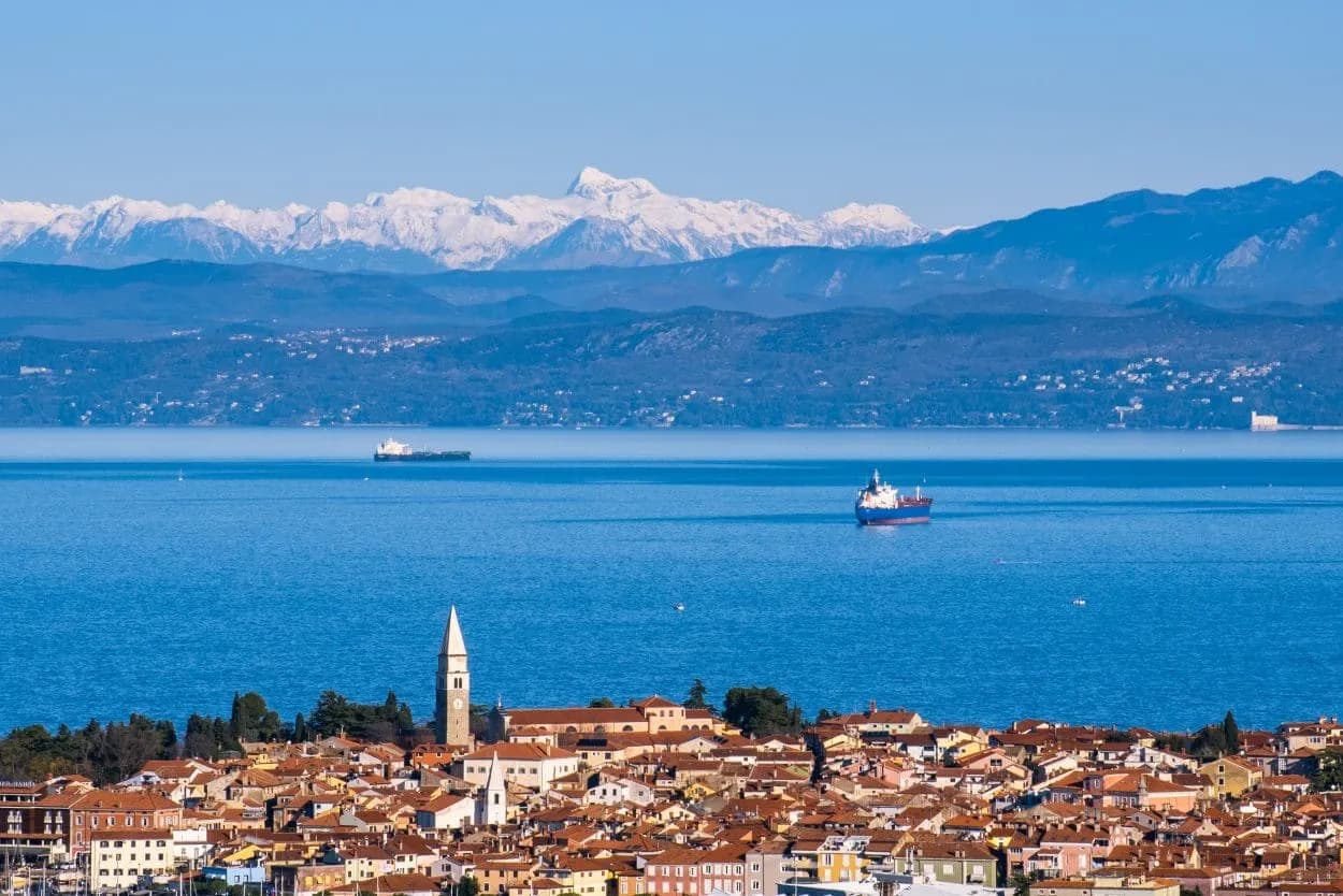 Coastal town with red roofs and bell tower, blue sea, and snow-capped mountains in background.