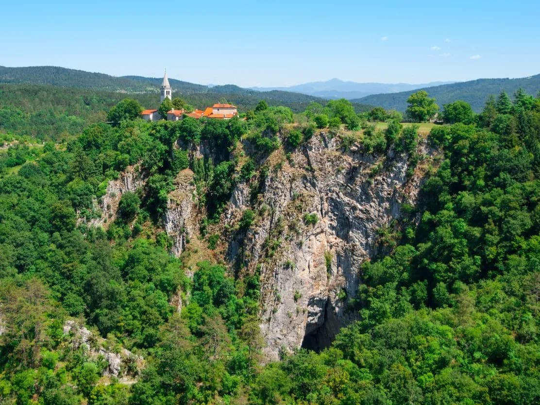 Village with church atop a steep, forested cliff above the entrance to Skocjan Caves.