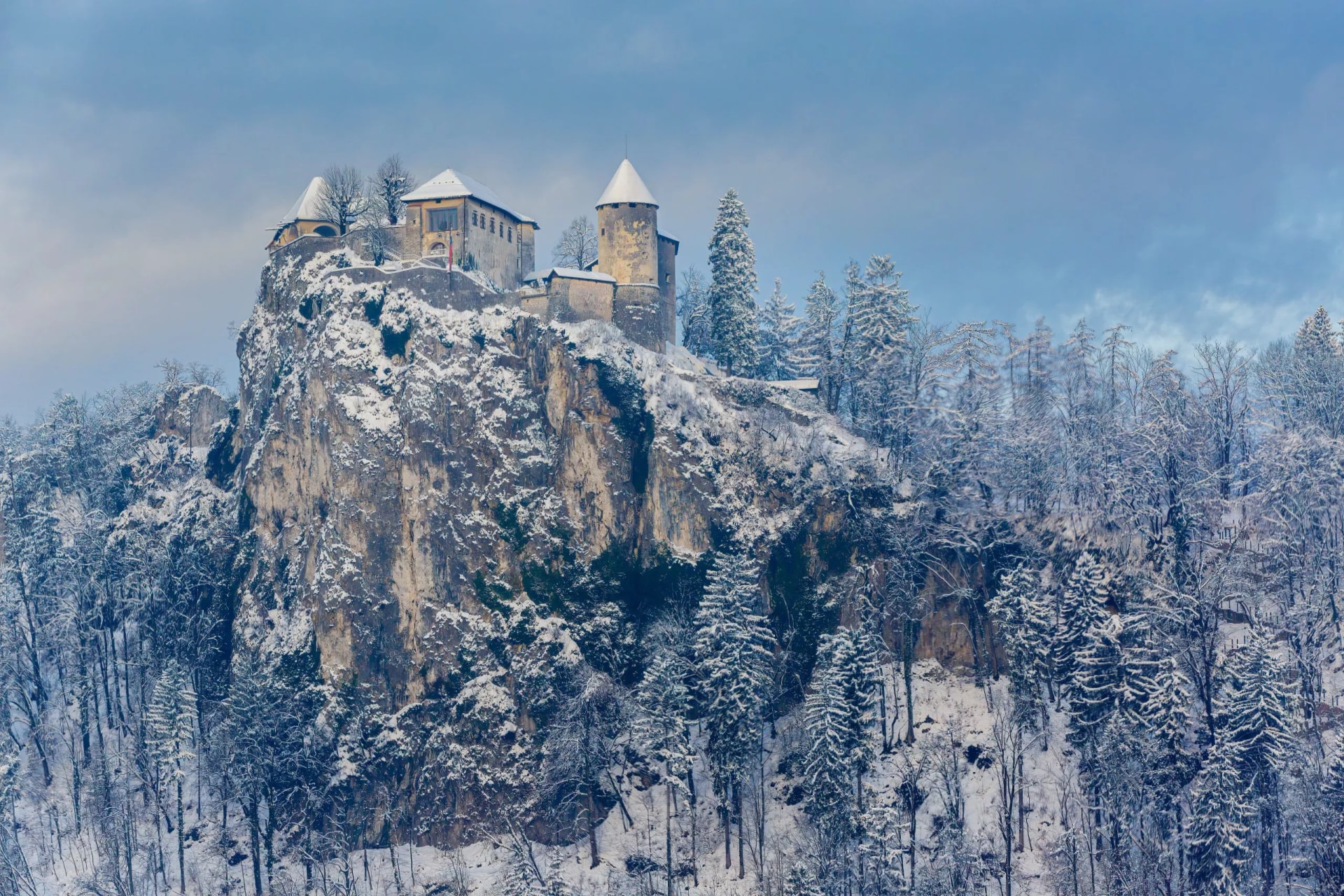 Bled Castle on a snow-covered cliff above winter forest under a cloudy blue sky.