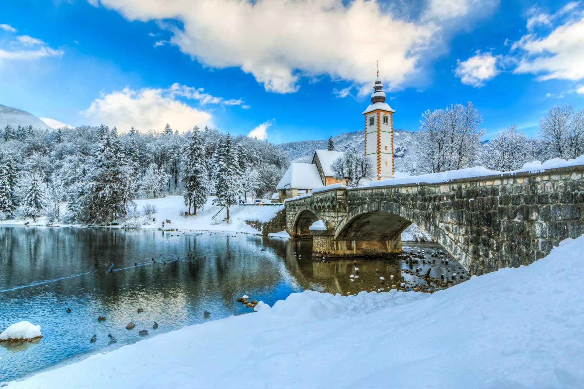 Stone bridge and church tower by Lake Bohinj in winter with snow-covered trees and blue sky.