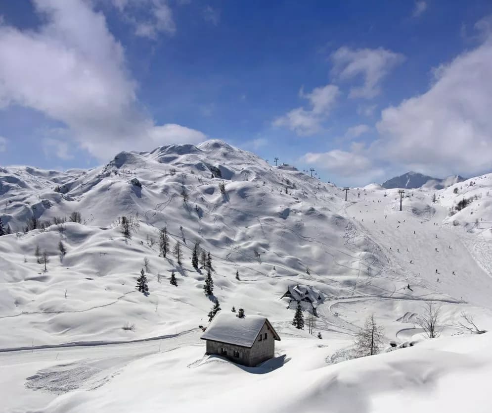 Small wooden cabin in deep snow at a ski resort with blue sky and snow-covered mountains.