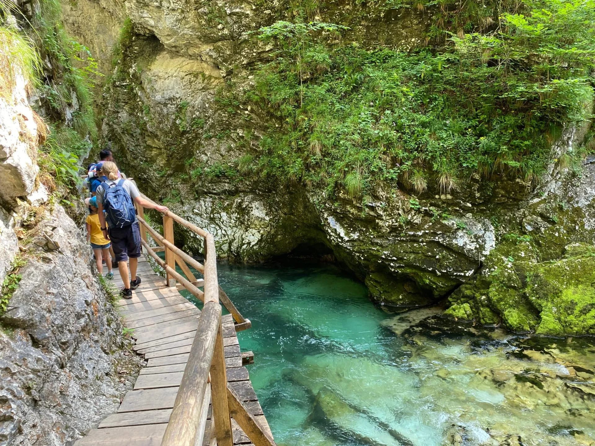 Hikers on wooden walkway alongside turquoise water in Vintgar Gorge canyon.
