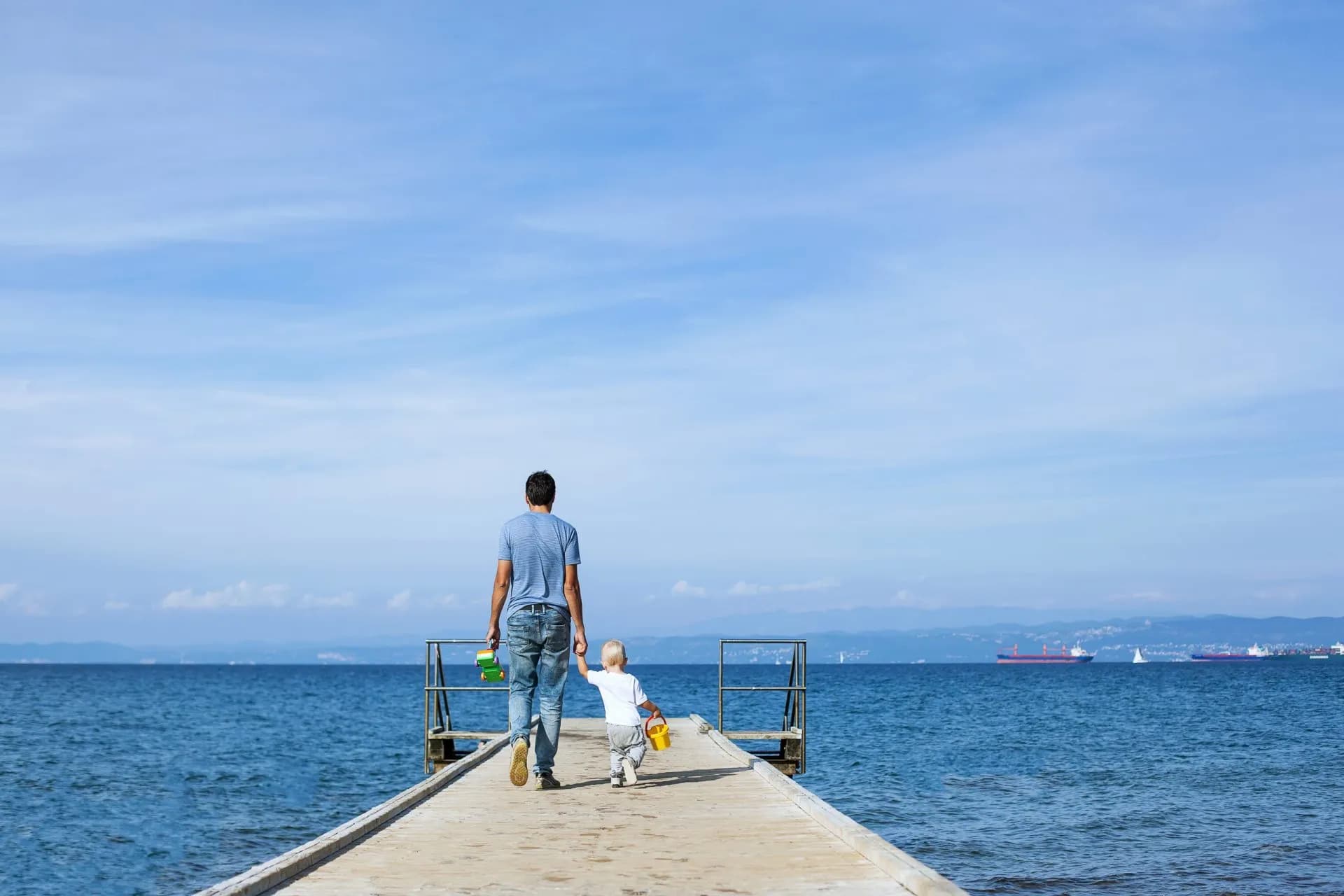 Father and child walk on wooden pier over blue sea toward distant coastline with ships.