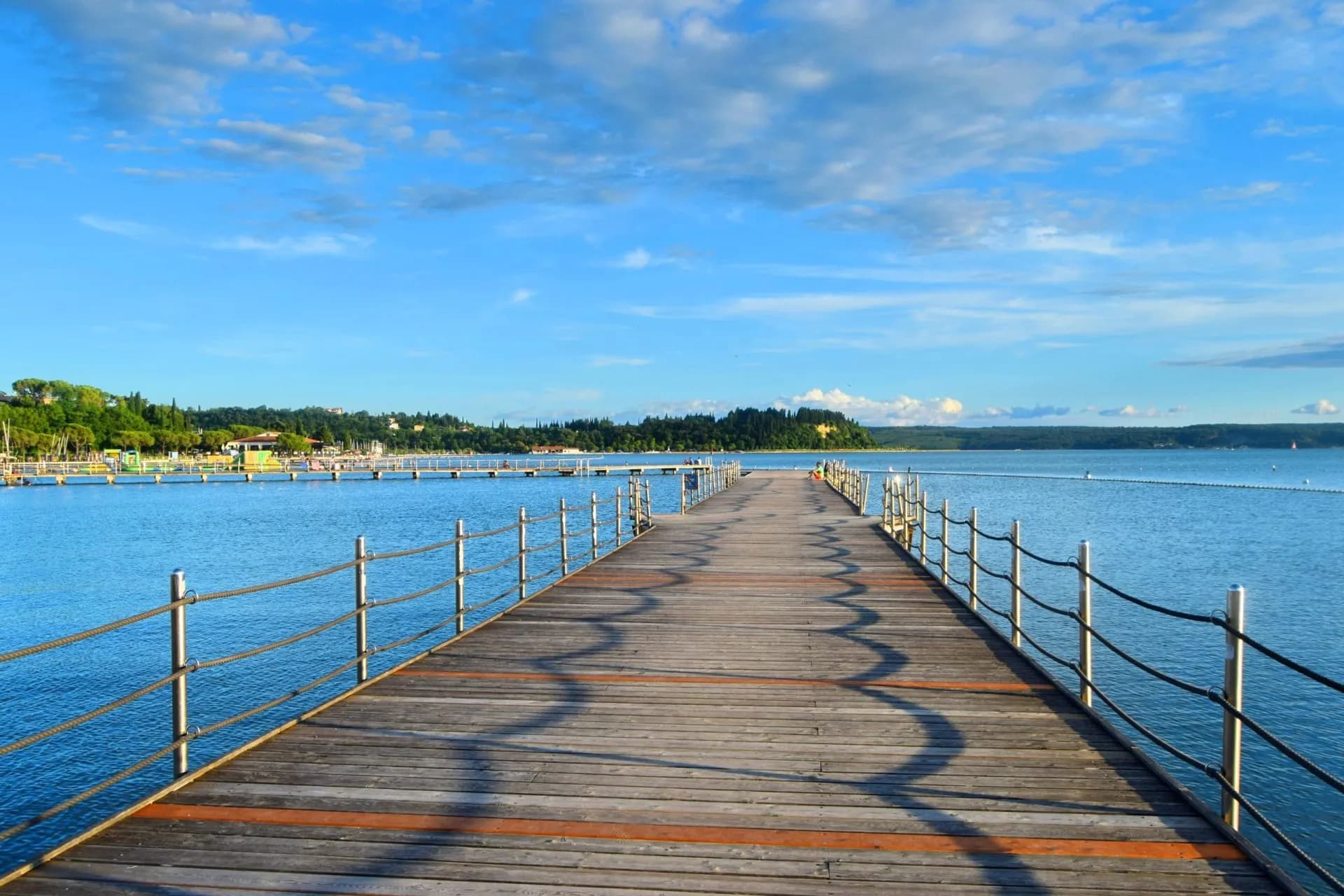 Wooden pier extending over blue sea toward a forested coastline under a blue sky.