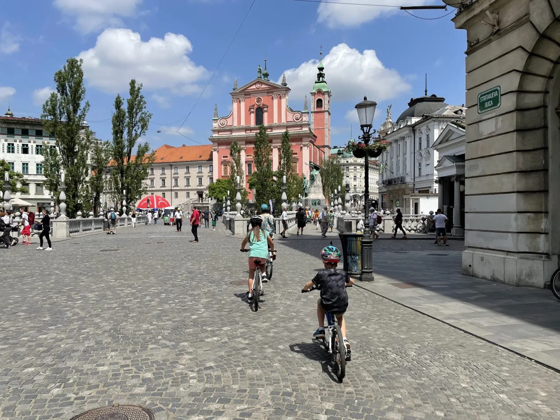 Children cycling on cobblestones toward the pink Franciscan Church in Ljubljana.