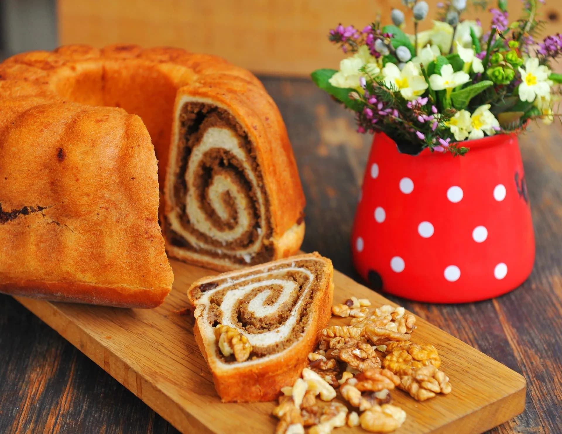 Walnut swirl bread (Potica) sliced on a wooden board with walnuts and flowers in a red vase.