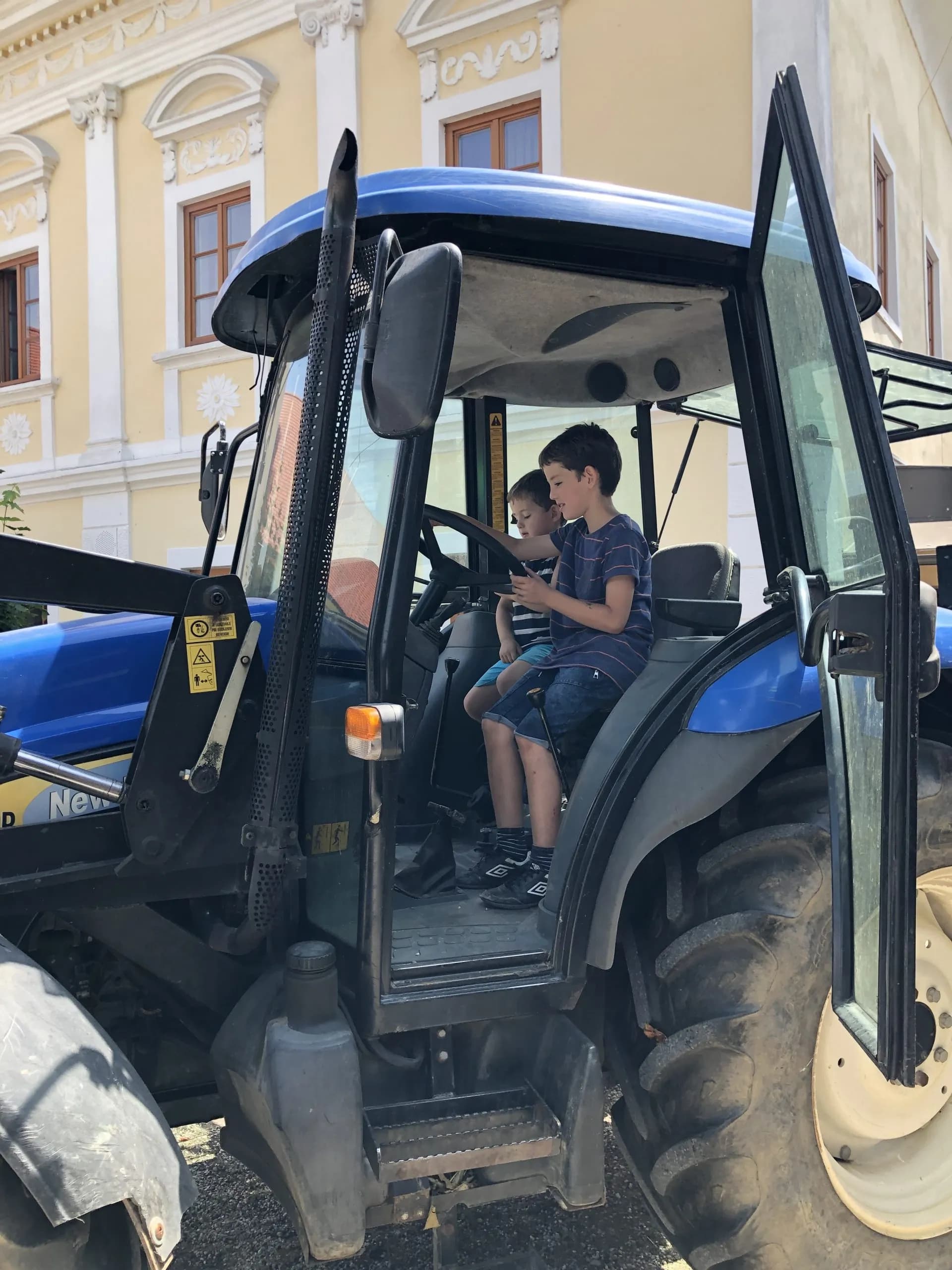 Two boys sitting inside a blue tractor parked near a yellow building with white trim.