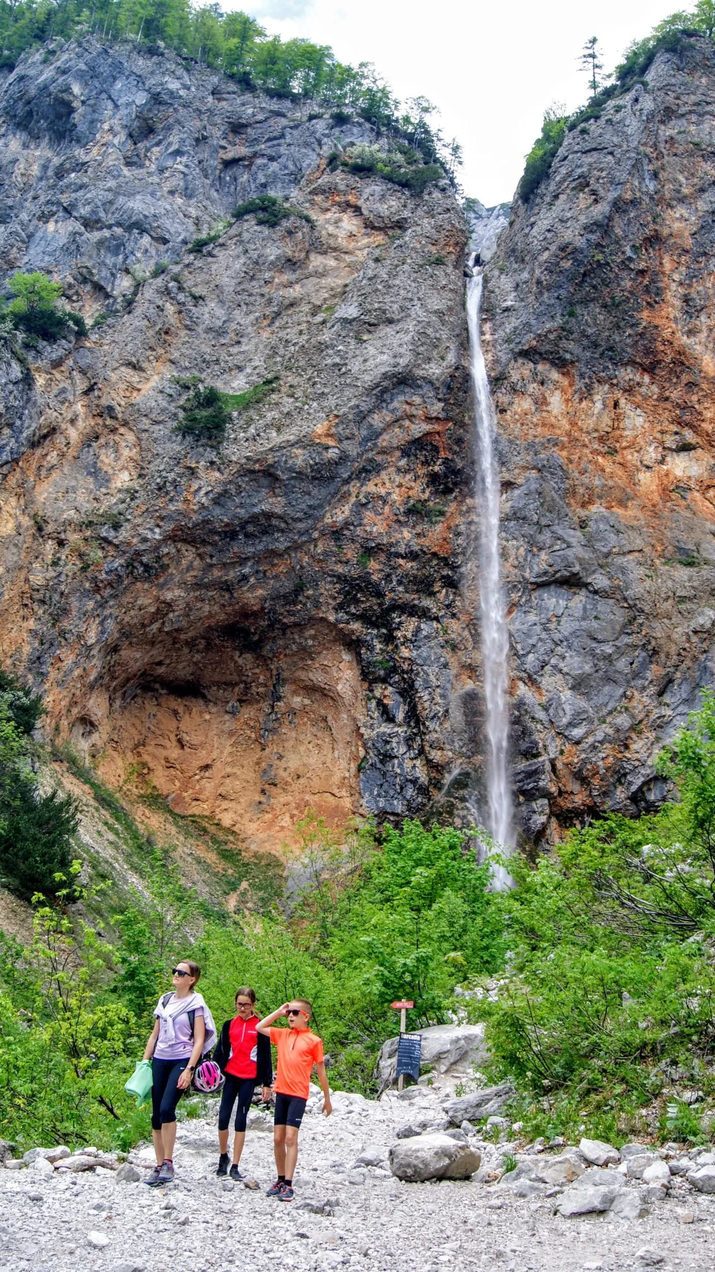 Hikers near a tall waterfall cascading down a rugged, colorful cliff face.